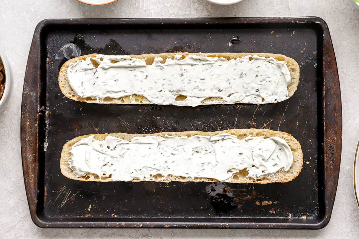 Toasted baguette halves spread with a thick layer of garlic herb cream cheese on a baking sheet.