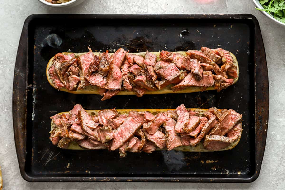 Sliced cooked steak piled onto the bottom half of toasted baguettes, ready to be assembled into sandwiches.
