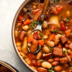 A spoon hovers over a hearty bowl of 15 bean soup, showcasing tender beans, diced carrots, smoked sausage, and fresh herbs in a rich, tomato-based broth.