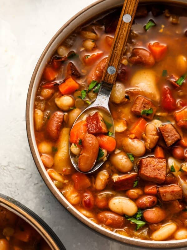 A spoon hovers over a hearty bowl of 15 bean soup, showcasing tender beans, diced carrots, smoked sausage, and fresh herbs in a rich, tomato-based broth.