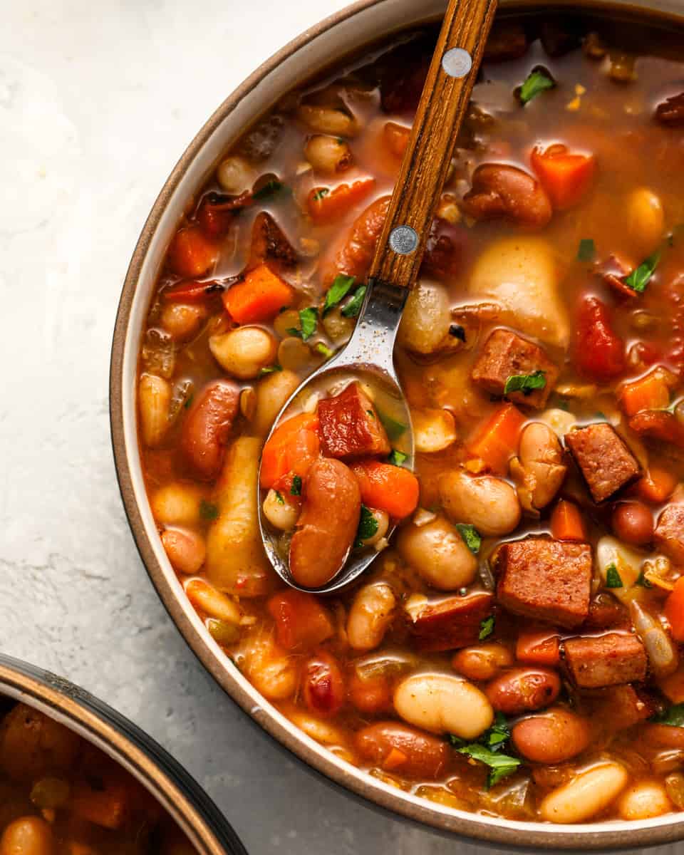 A spoon hovers over a hearty bowl of 15 bean soup, showcasing tender beans, diced carrots, smoked sausage, and fresh herbs in a rich, tomato-based broth.