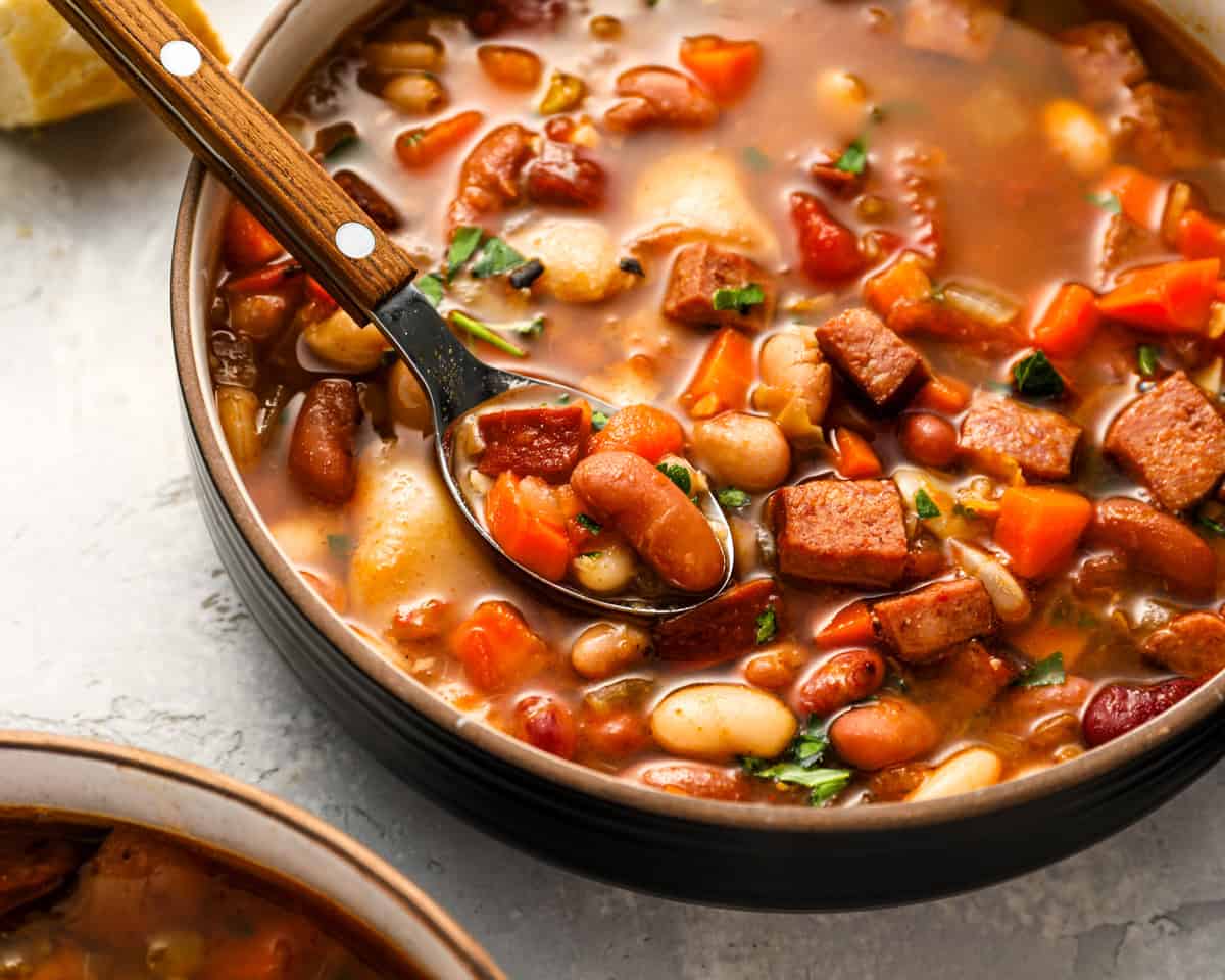 A bowl of colorful 15 bean soup with a spoon lifting a scoop of beans, sausage, and vegetables; garnished with parsley and surrounded by lemon wedges.