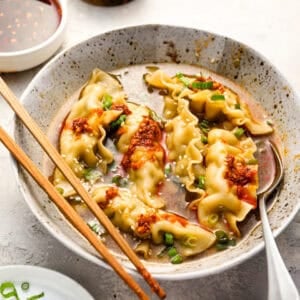 A speckled ceramic bowl filled with dumpling soup, topped with chili crisp, chopped scallions, and sesame seeds, with chopsticks and a spoon resting on the side.