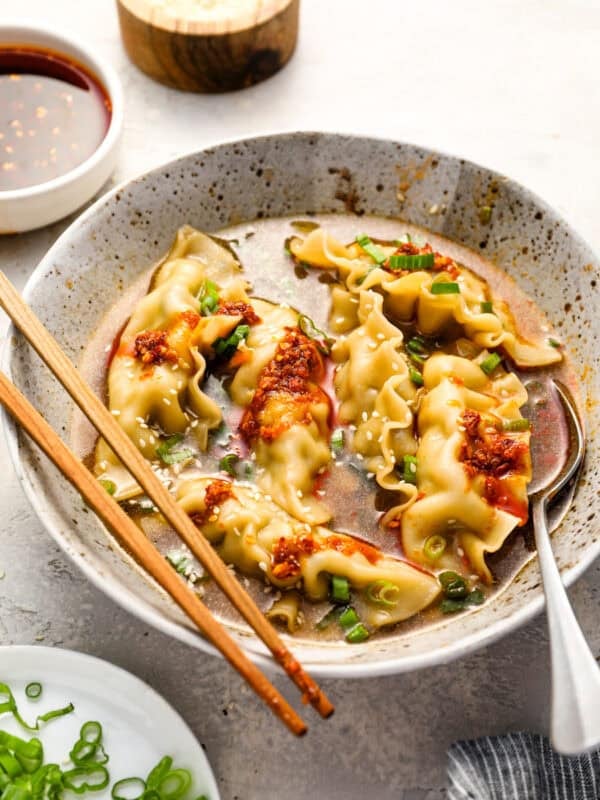 A speckled ceramic bowl filled with dumpling soup, topped with chili crisp, chopped scallions, and sesame seeds, with chopsticks and a spoon resting on the side.