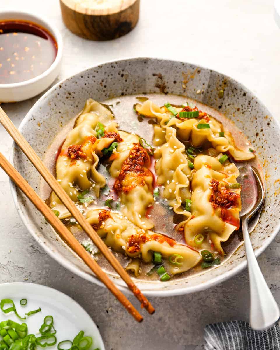 A speckled ceramic bowl filled with dumpling soup, topped with chili crisp, chopped scallions, and sesame seeds, with chopsticks and a spoon resting on the side.