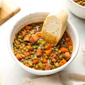 A close-up of a bowl of ham and lentil soup with diced carrots, celery, and ham, served with a piece of crusty bread.