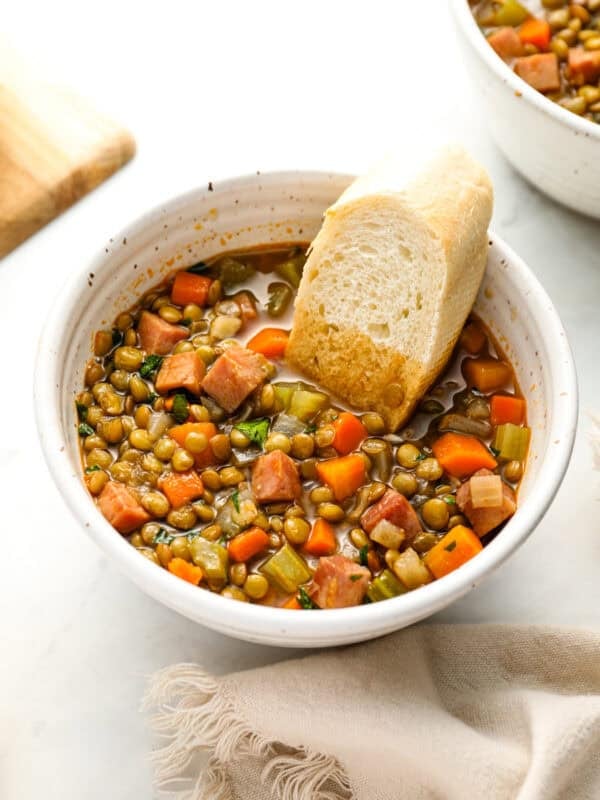 A close-up of a bowl of ham and lentil soup with diced carrots, celery, and ham, served with a piece of crusty bread.