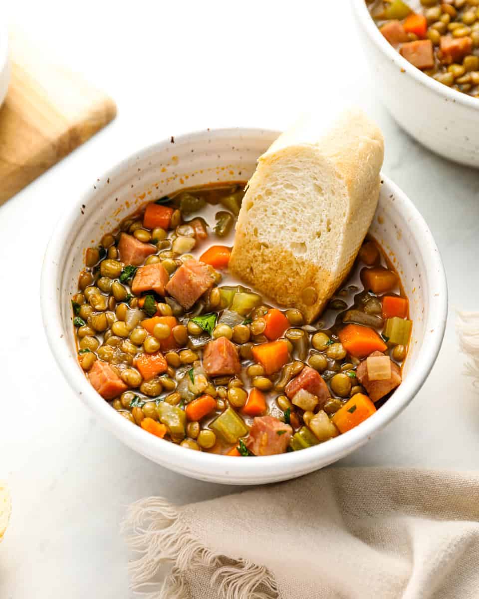  A close-up of a bowl of ham and lentil soup with diced carrots, celery, and ham, served with a piece of crusty bread.
