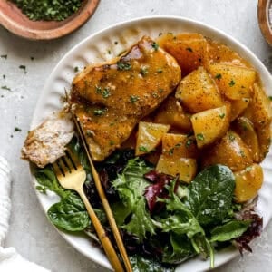 A white plate filled with creamy crockpot pork chops, seasoned potatoes, and a side of fresh mixed greens, served with gold flatware.
