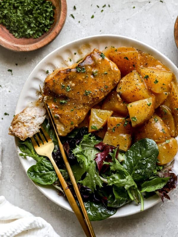 A white plate filled with creamy crockpot pork chops, seasoned potatoes, and a side of fresh mixed greens, served with gold flatware.
