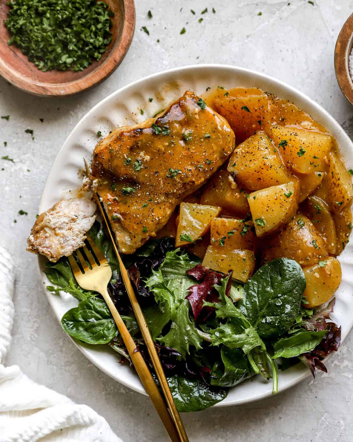 A white plate filled with creamy crockpot pork chops, seasoned potatoes, and a side of fresh mixed greens, served with gold flatware.