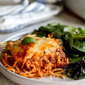 A plated slice of baked million dollar spaghetti topped with melted cheese and fresh basil, served alongside a simple green salad.
