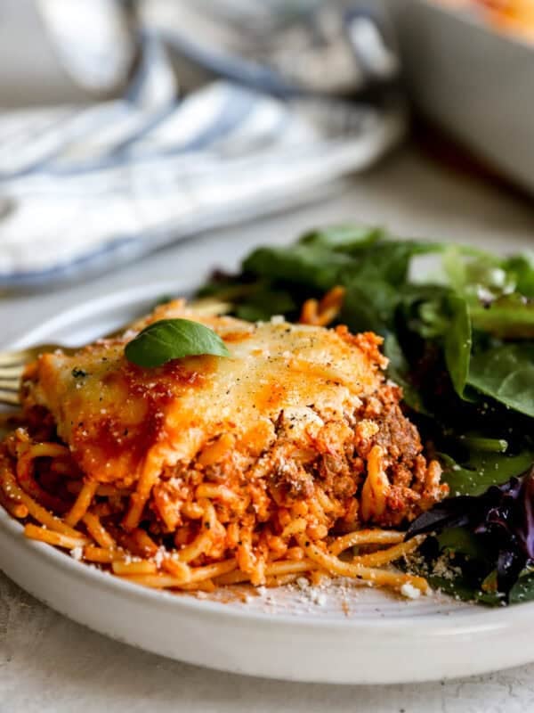 A plated slice of baked million dollar spaghetti topped with melted cheese and fresh basil, served alongside a simple green salad.