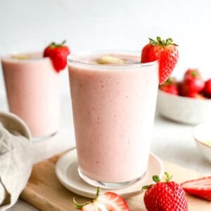 Close-up of a strawberry banana smoothie in a glass garnished with a fresh strawberry and banana slice, with a second smoothie in the background and strawberries scattered on a cutting board.
