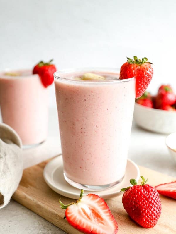 Close-up of a strawberry banana smoothie in a glass garnished with a fresh strawberry and banana slice, with a second smoothie in the background and strawberries scattered on a cutting board.