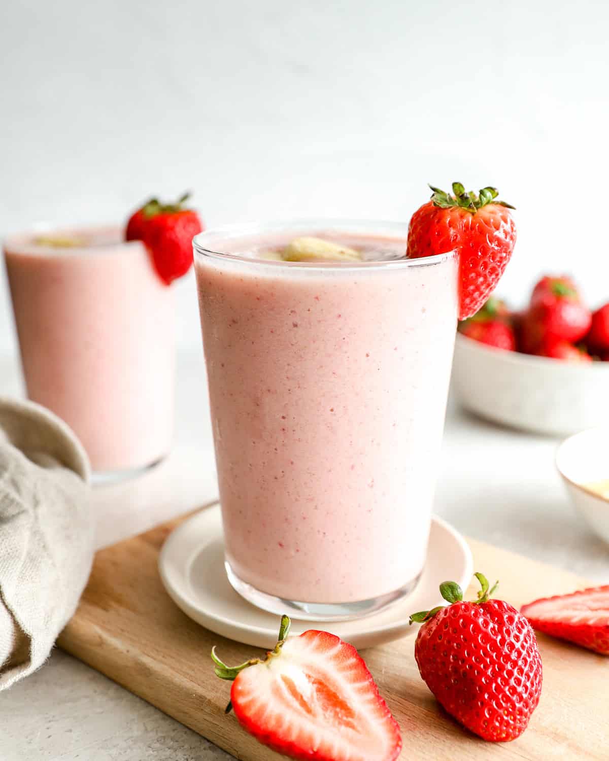 Close-up of a strawberry banana smoothie in a glass garnished with a fresh strawberry and banana slice, with a second smoothie in the background and strawberries scattered on a cutting board.