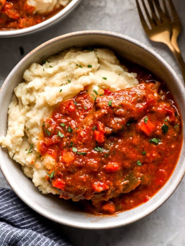 Bowl of Swiss steak served over mashed potatoes, topped with rich tomato-based sauce and sprinkled with chopped parsley.