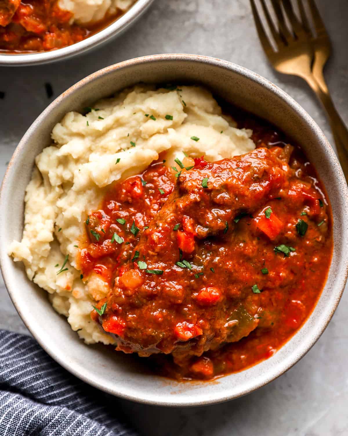 Bowl of Swiss steak served over mashed potatoes, topped with rich tomato-based sauce and sprinkled with chopped parsley.