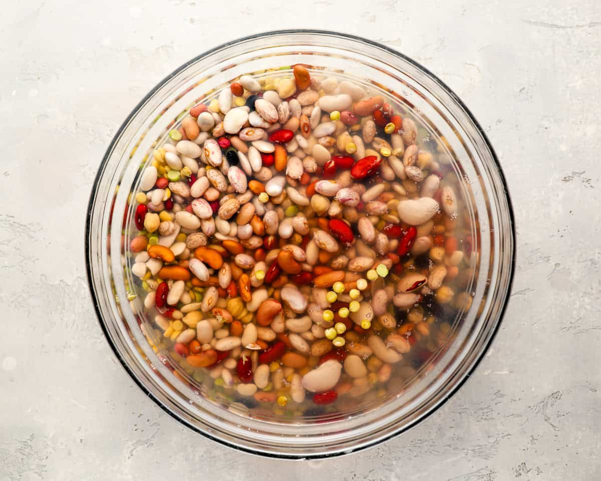 A bowl of dried 15 bean mix soaking in water, with a variety of colorful beans like kidney, lima, and pinto fully visible.