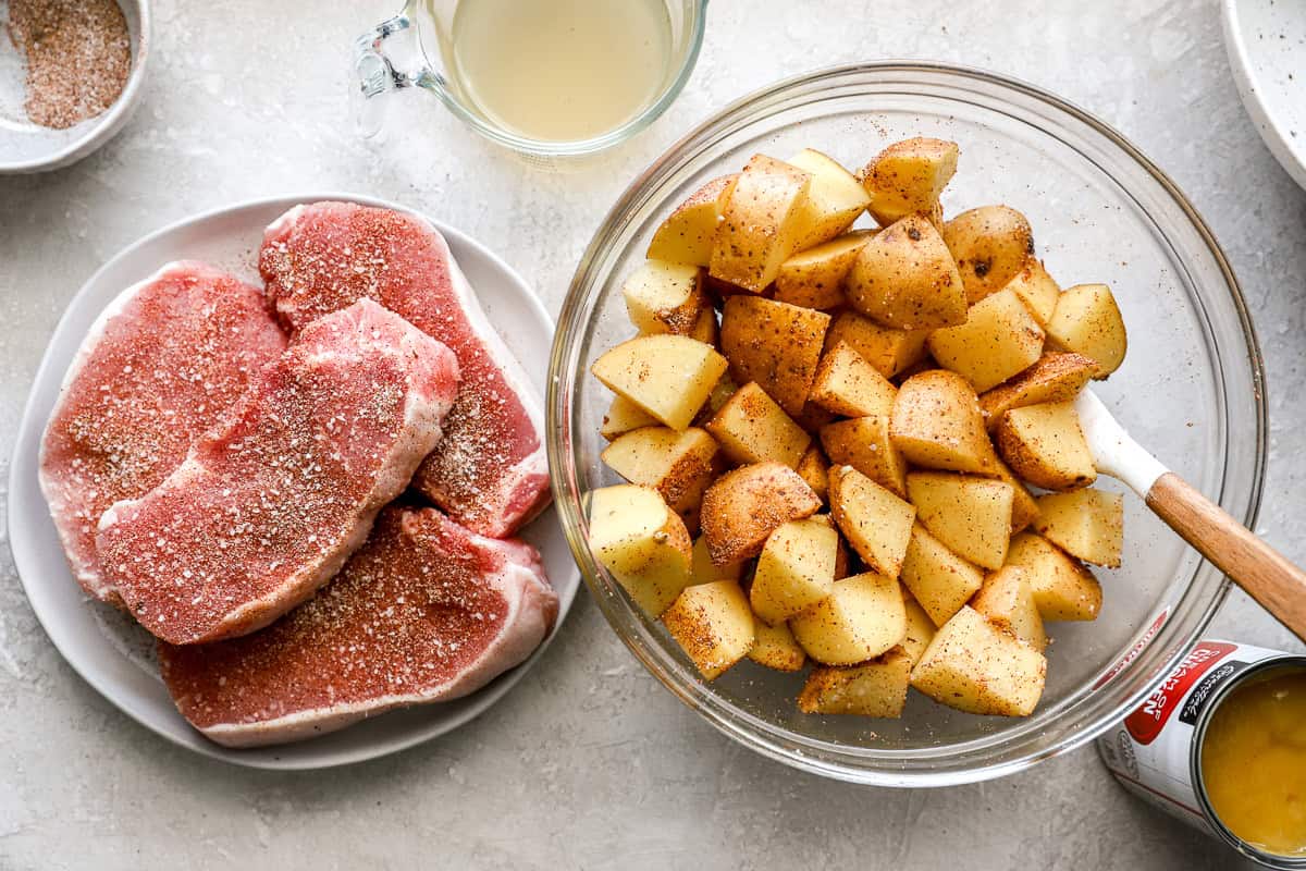 A prep scene with raw pork chops on a plate, seasoned potato chunks in a glass bowl, chicken broth in a measuring cup, and a can of condensed soup nearby.