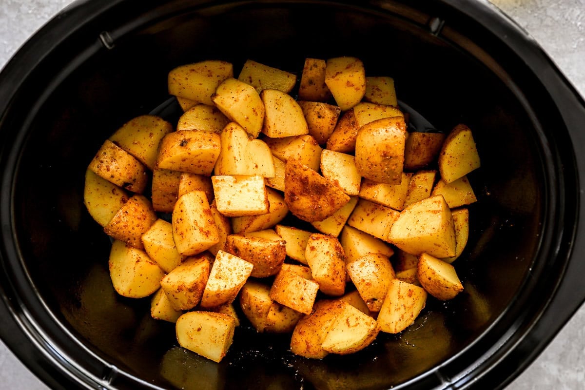Seasoned potato chunks spread in an even layer inside a black crockpot, ready for slow cooking.