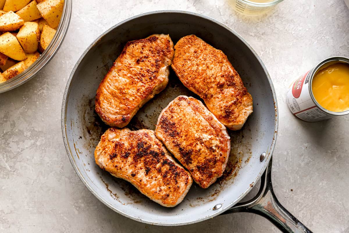 Four pork chops searing in a skillet with golden brown edges, surrounded by prep ingredients like potatoes and soup.