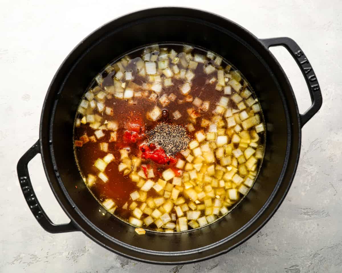 An overhead shot of a Dutch oven filled with broth, diced onions, black pepper, and tomatoes before adding the beans and sausage.