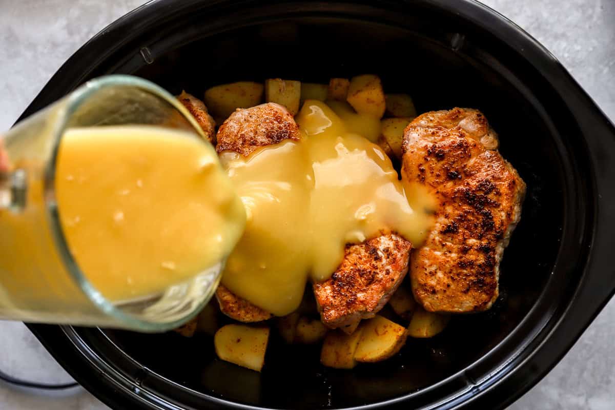 Creamy soup mixture being poured from a glass measuring cup over seared pork chops in a crockpot layered on top of potatoes.