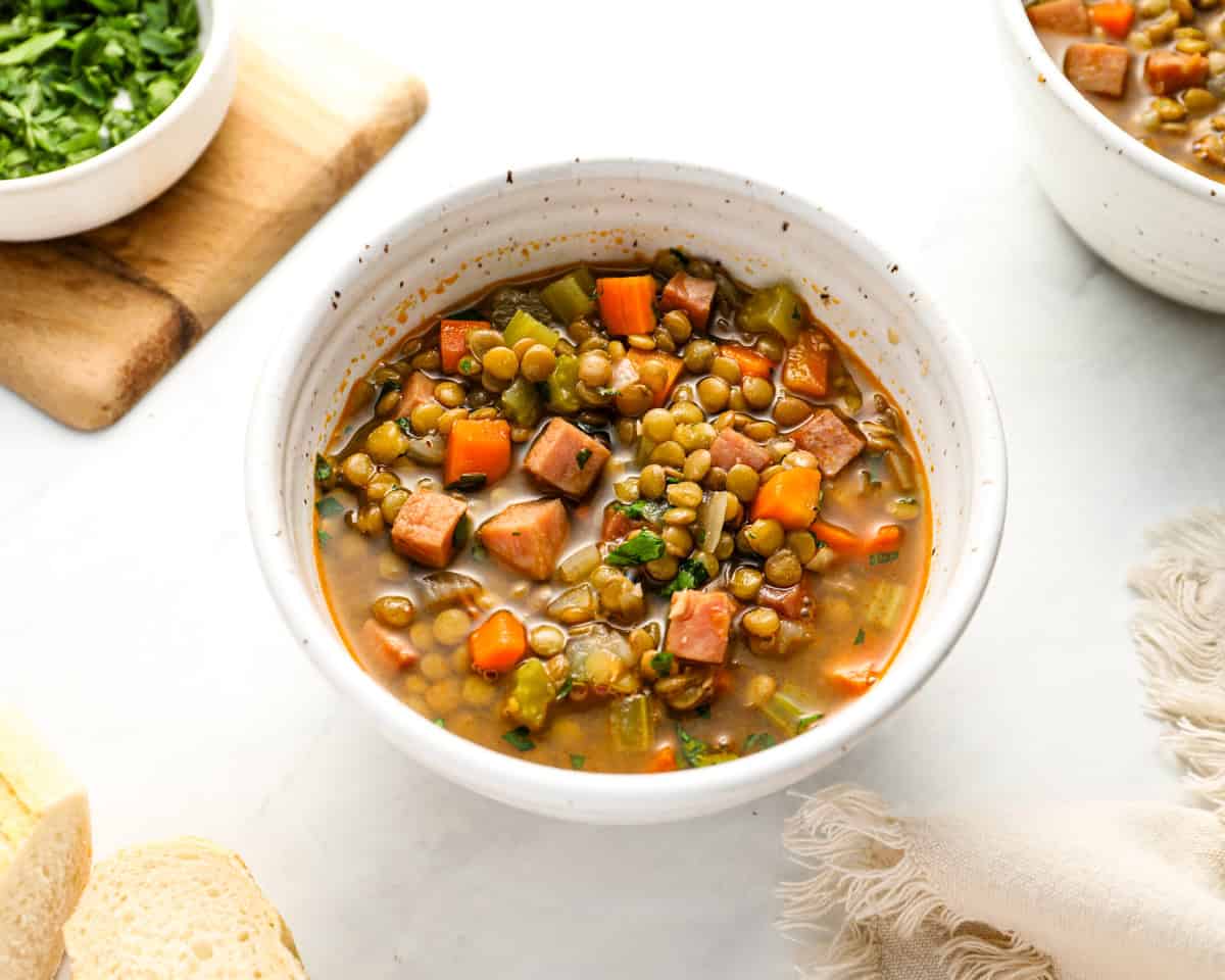 A bowl of ham and lentil soup on a white surface, garnished with parsley and served alongside another bowl and fresh bread.