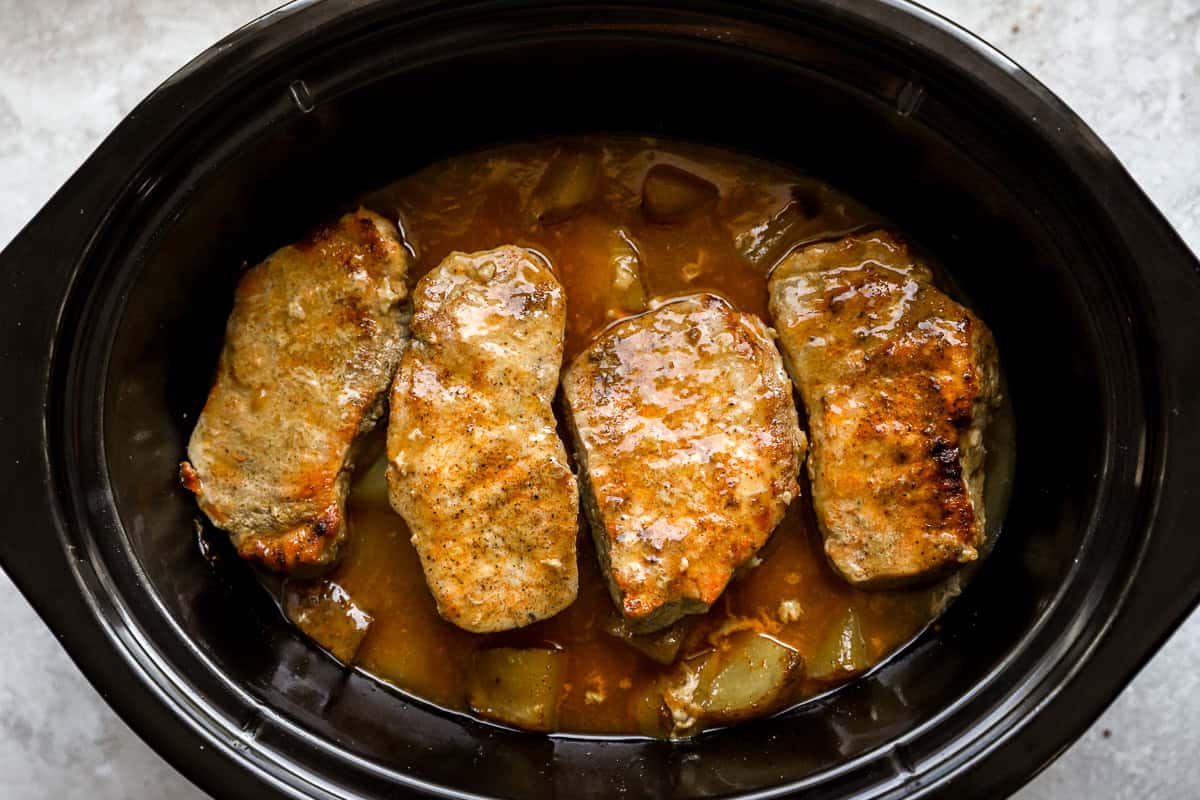 Four golden, tender pork chops sitting in a rich, creamy sauce over potatoes in a crockpot after cooking.
