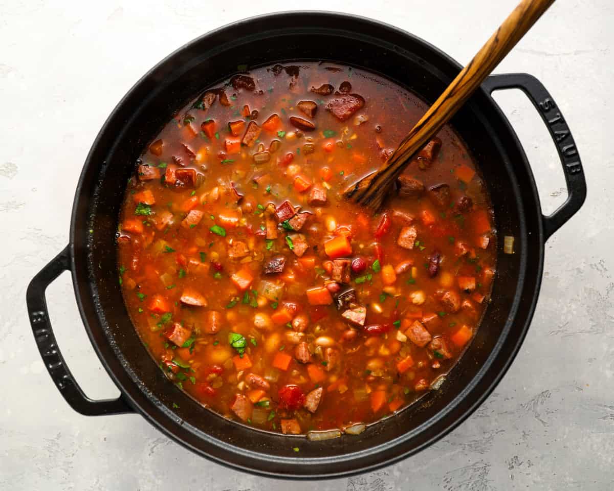 A large black Dutch oven filled with simmering 15 bean soup, with a wooden spoon resting inside and the soup dotted with fresh herbs.