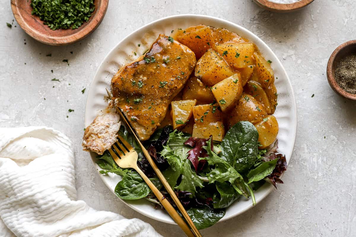 Close-up of a serving of crockpot pork chops and potatoes on a white plate with mixed greens and a forkful of pork.