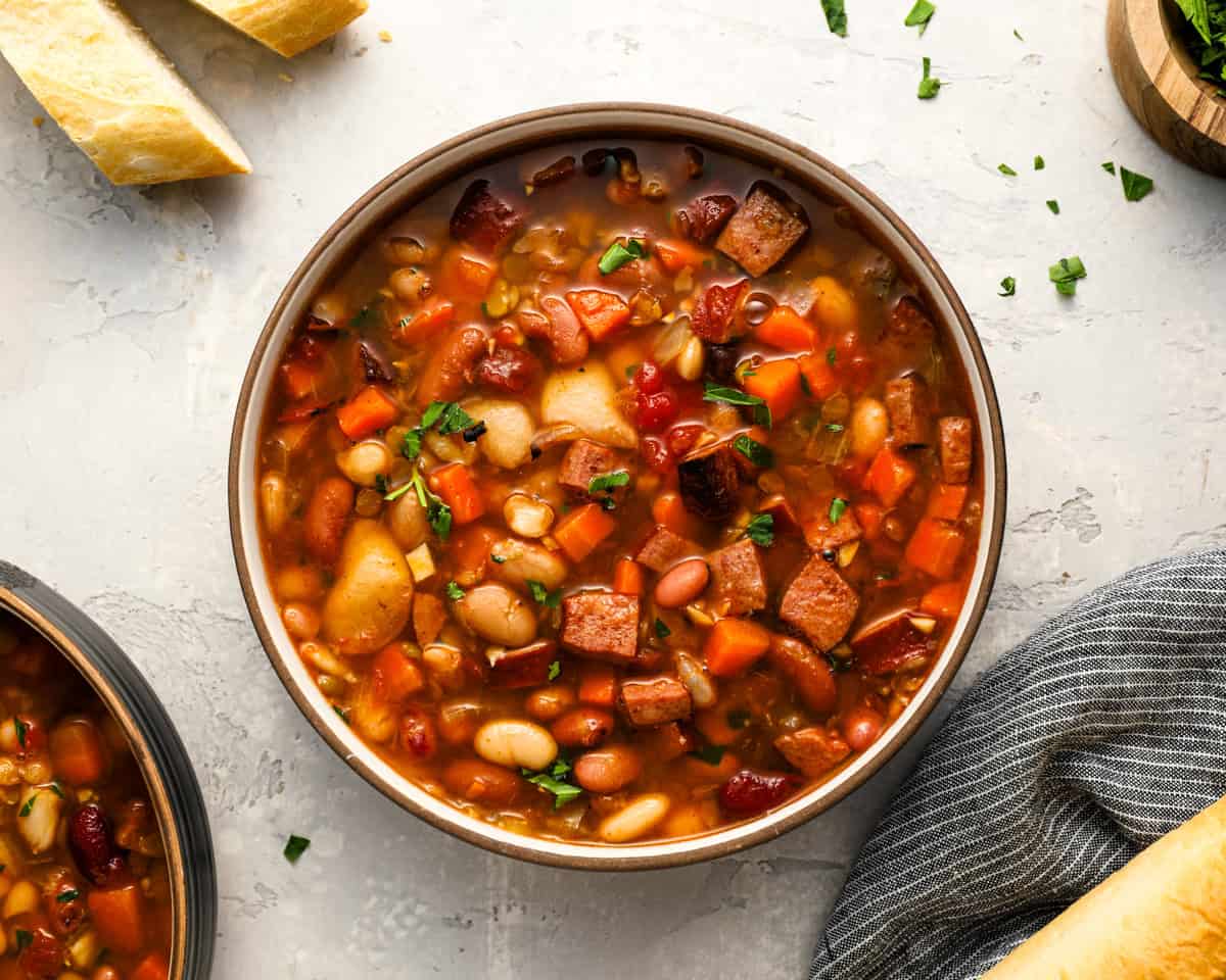 A top-down view of a full bowl of 15 bean soup with visible chunks of sausage, carrots, and beans, sprinkled with chopped parsley and served with bread on the side.