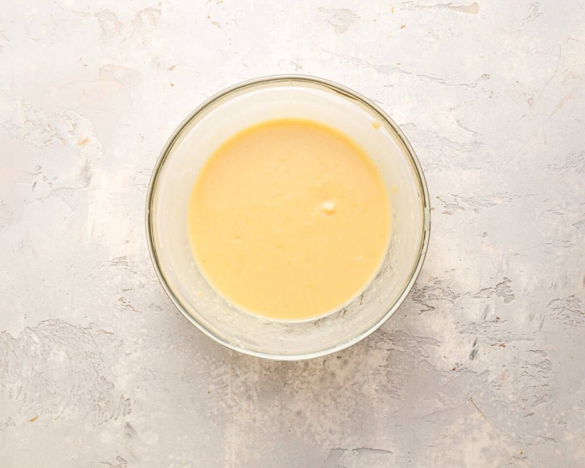 A glass bowl with a light yellow miso broth mixture on a textured white surface.