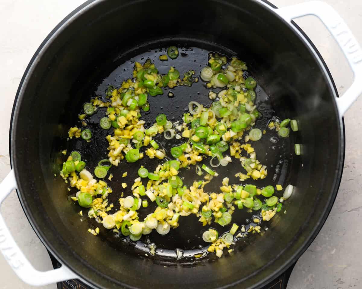 Aromatics including chopped green onions, garlic, and ginger sizzling in oil in a black Dutch oven.