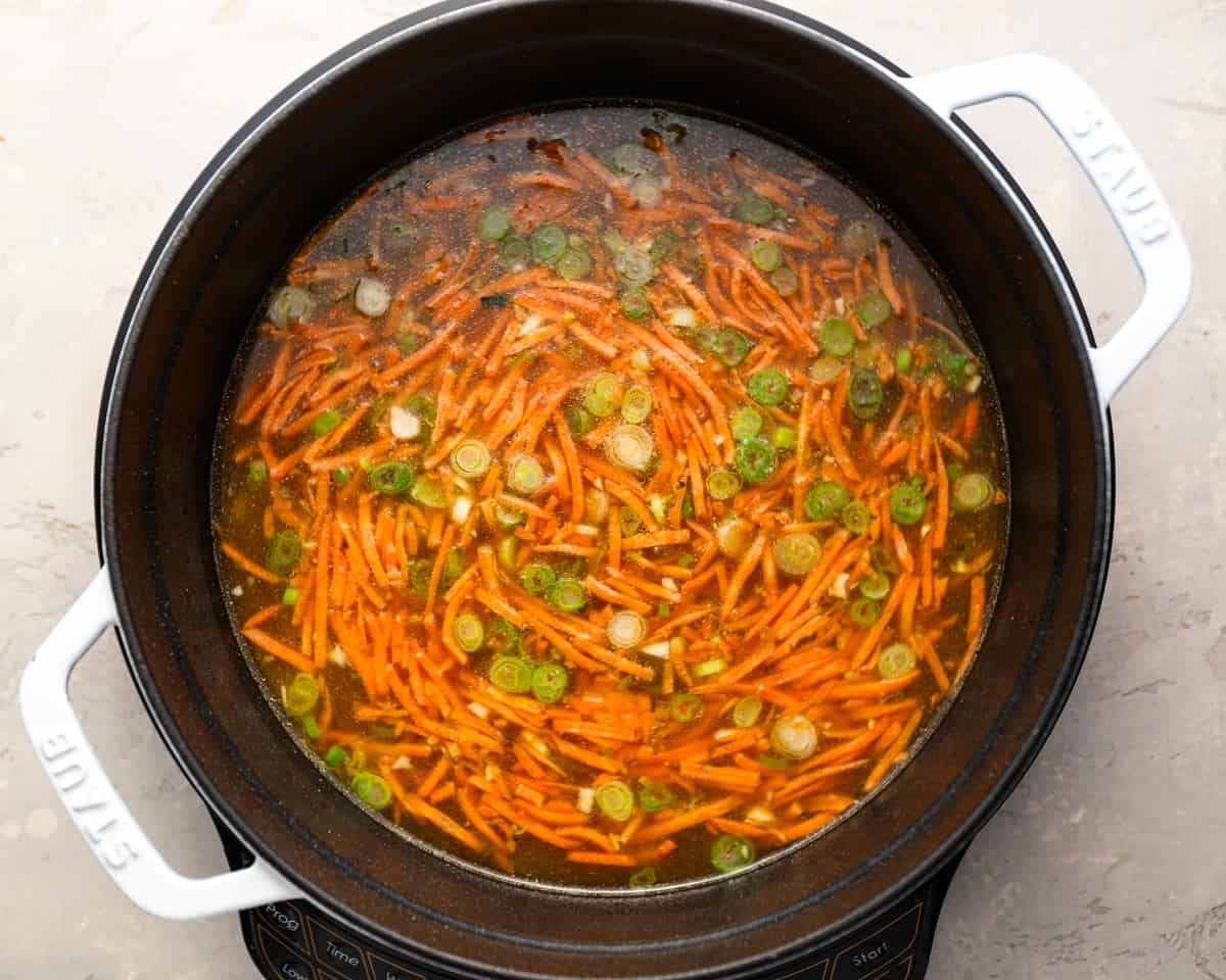 Shredded carrots and green onions simmering in broth inside a white Dutch oven.