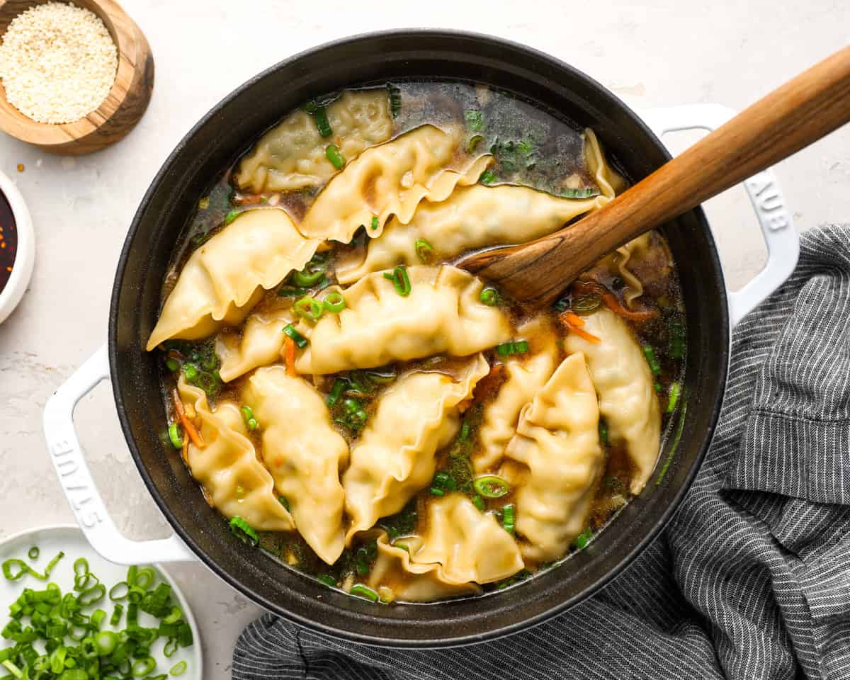 A wooden spoon stirring dumpling soup in a white Dutch oven, surrounded by bowls of sesame seeds and chili oil.