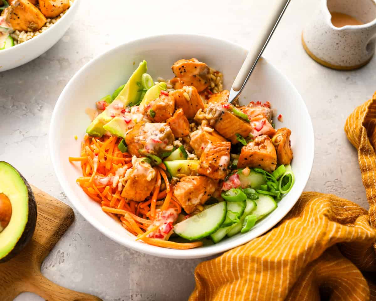 Close-up side view of a salmon rice bowl with avocado, cucumber, carrots, green onions, and sauce, with a fork ready to dig in.