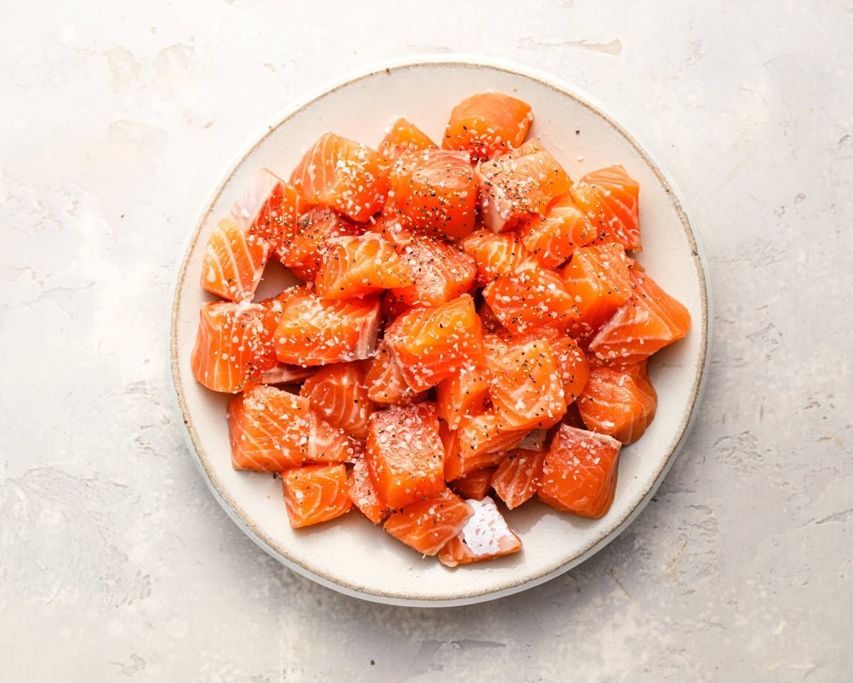 Raw cubed salmon seasoned with salt and pepper in a white bowl, ready to be cooked.