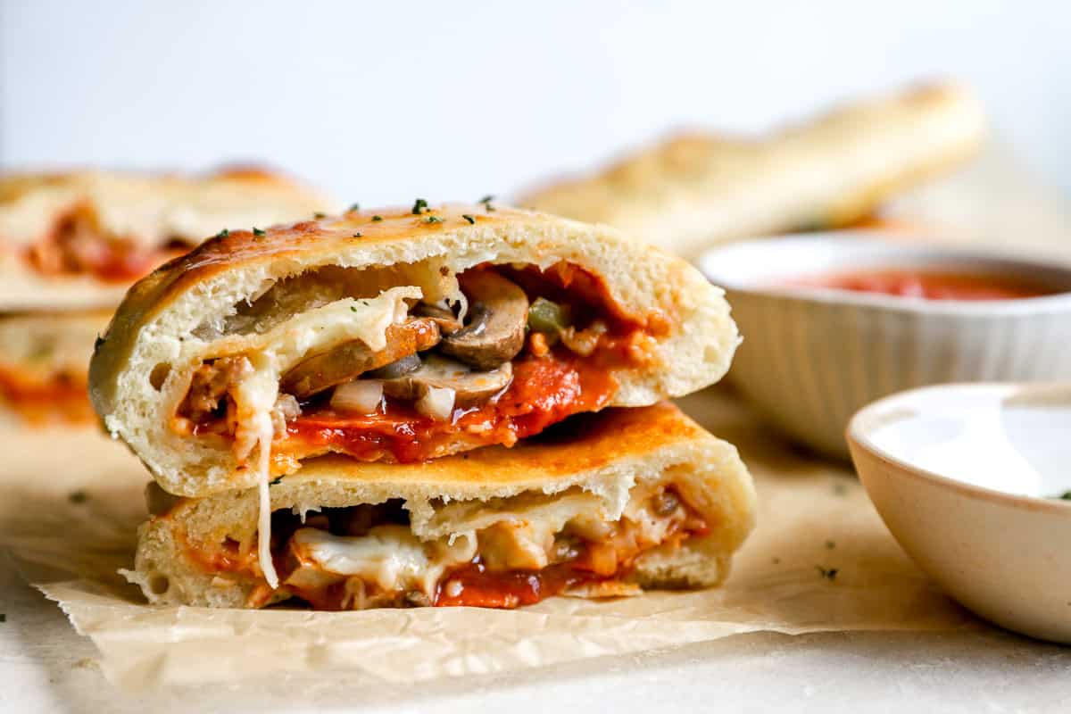 Close-up of sliced calzone showing melty cheese, pepperoni, mushrooms, and sauce inside, with dipping sauce in background.