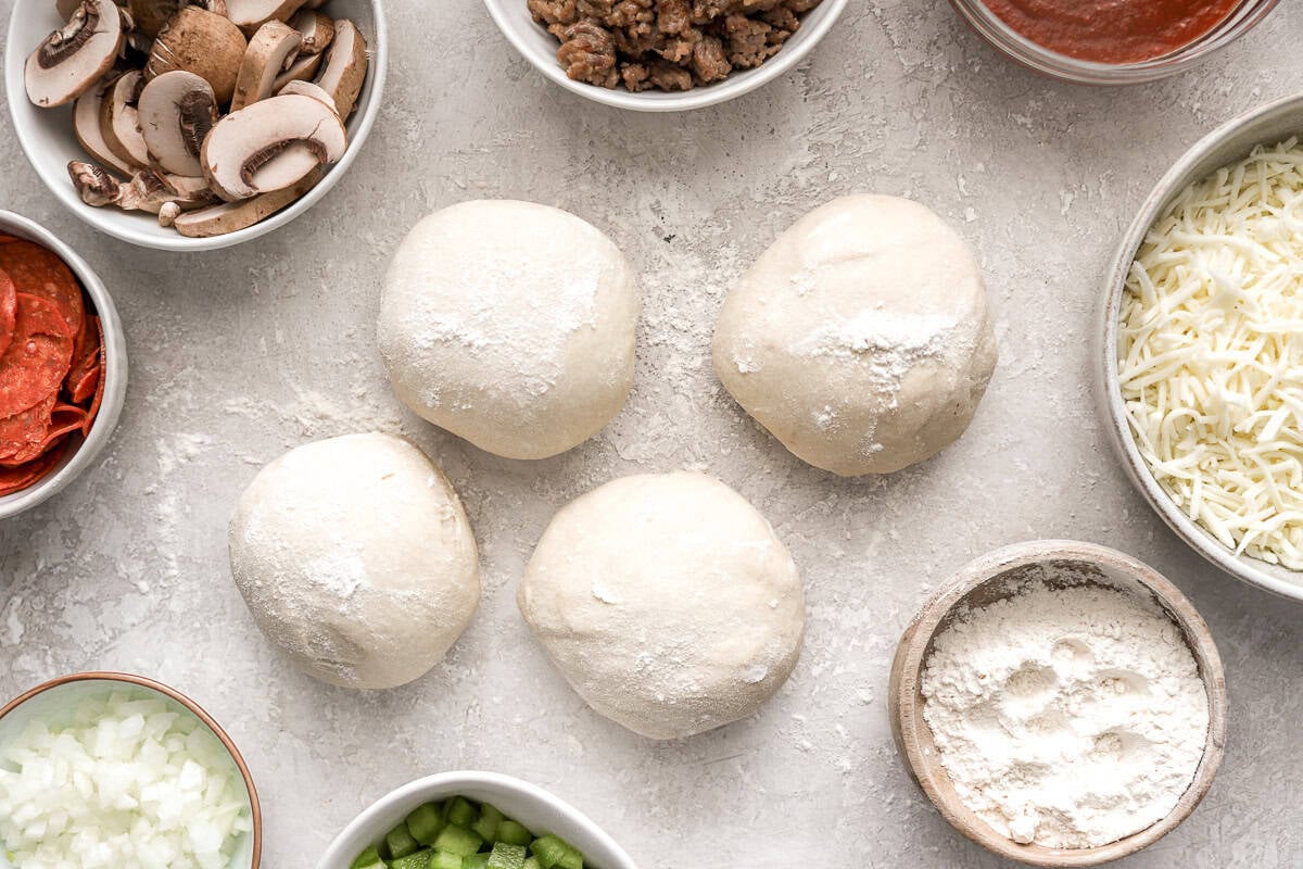 Ingredients laid out for making calzones, including dough balls, mushrooms, sausage, pizza sauce, cheese, pepperoni, and chopped vegetables.