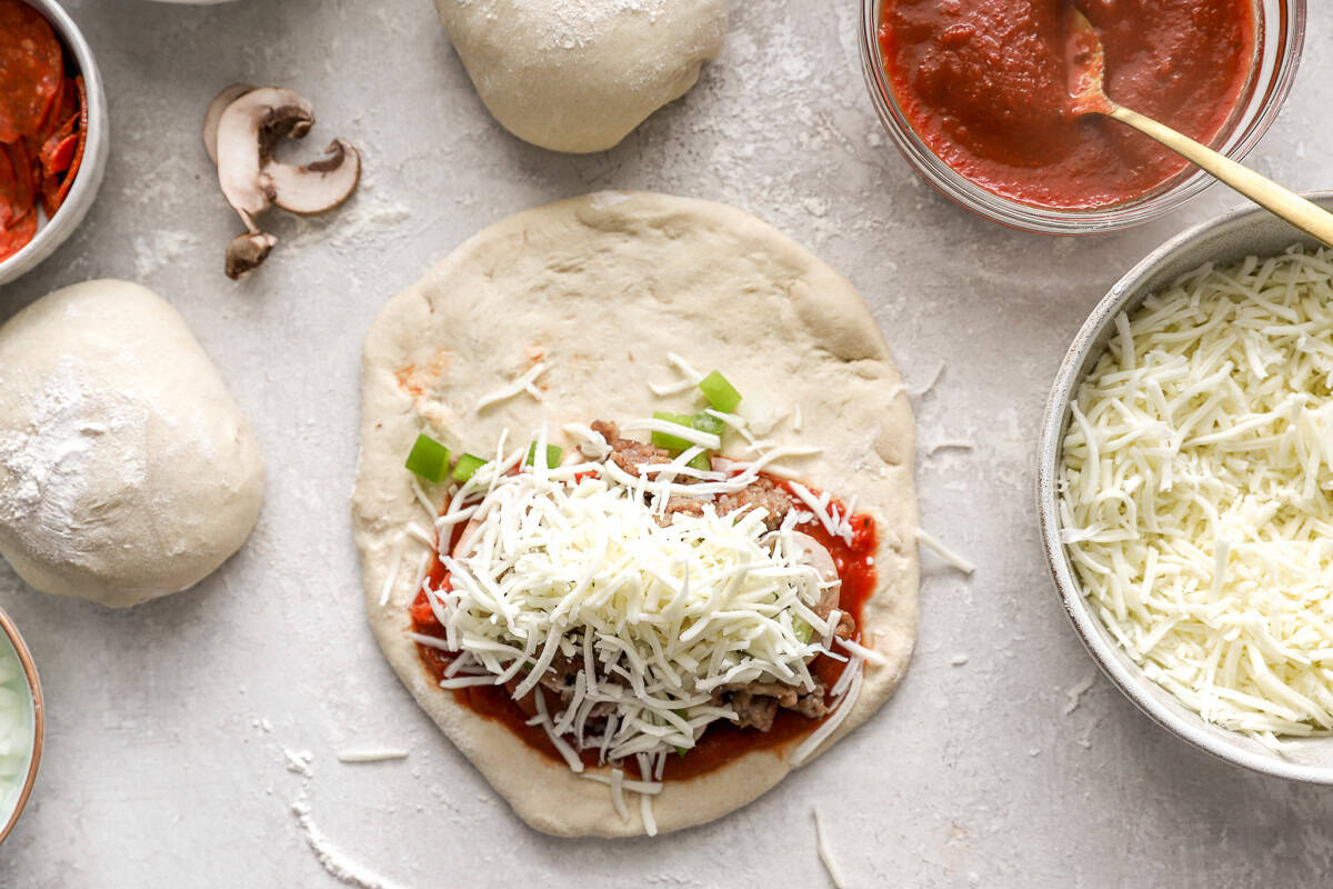 Calzone dough topped with pizza sauce, sausage, mushrooms, onions, bell peppers, and shredded mozzarella ready to be folded.