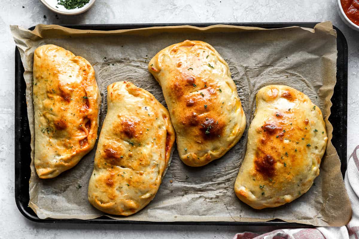 Four fully baked calzones on a baking sheet with golden, glossy crusts.