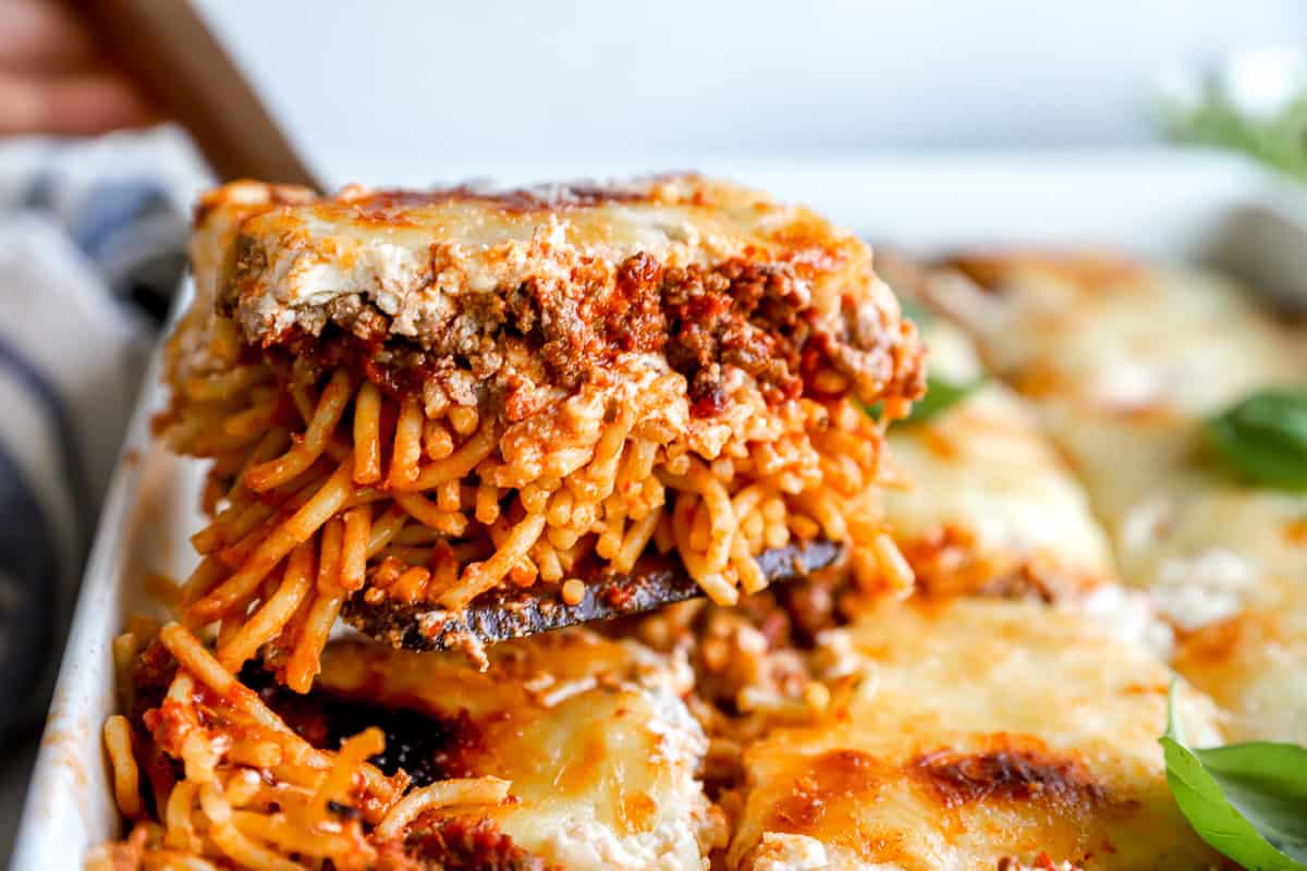 A spatula lifting a hearty slice of million dollar spaghetti, showing layers of noodles, meat sauce, creamy cheese, and melted mozzarella.