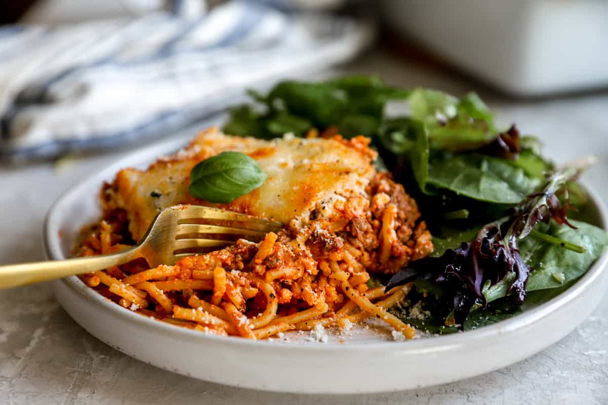 Close-up of a plated serving of million dollar spaghetti with a fork, garnished with basil and served next to fresh salad greens.