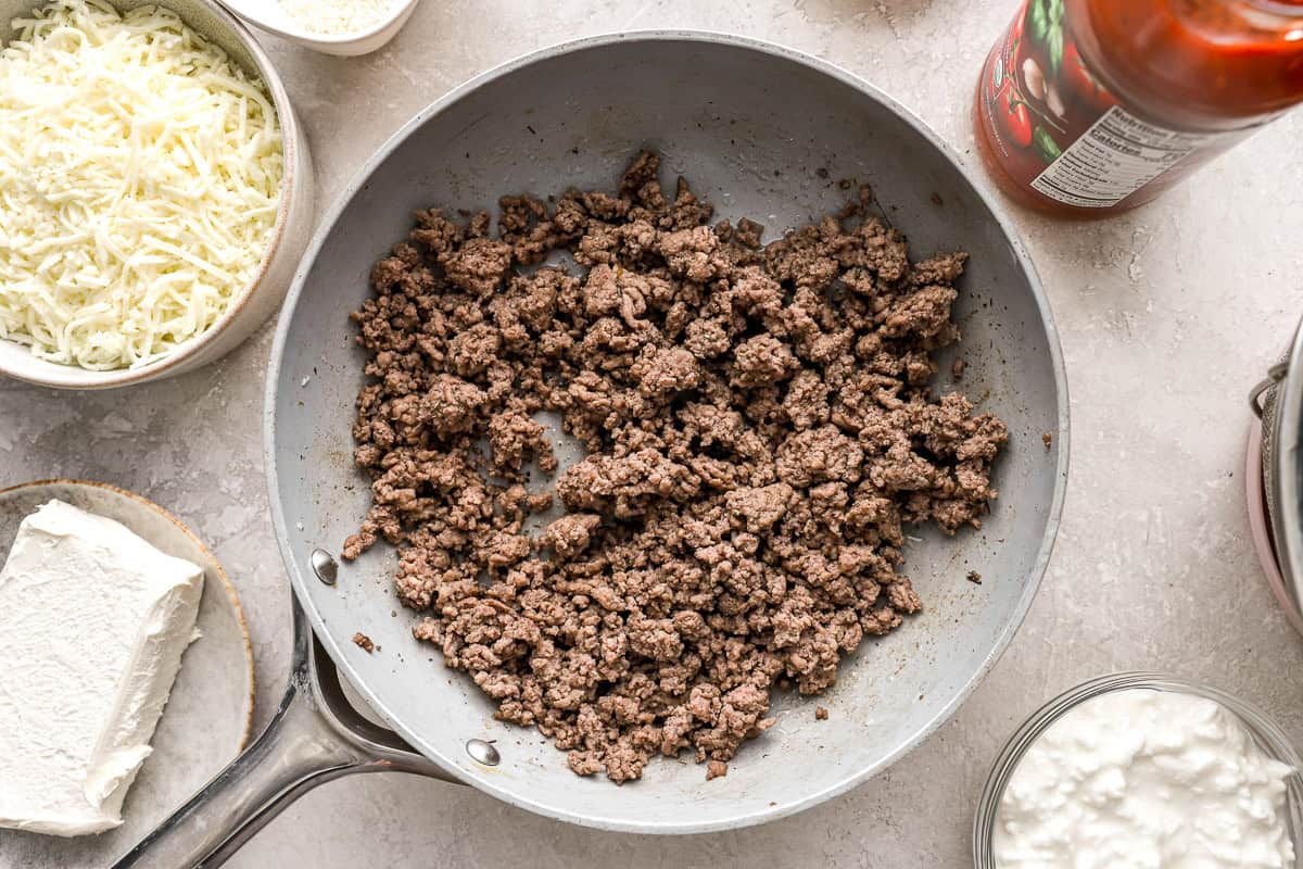 Cooked ground beef in a skillet surrounded by ingredients including shredded mozzarella, cream cheese, cottage cheese, and a jar of marinara sauce.