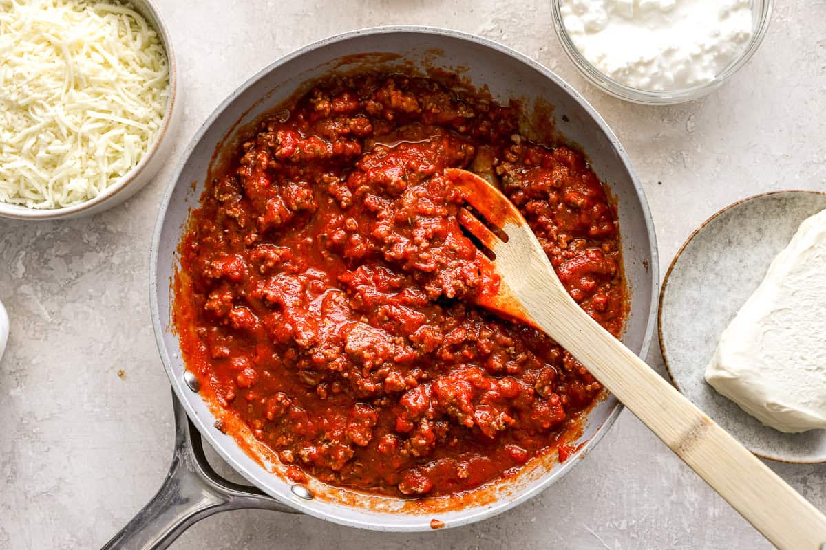 Ground beef simmering in marinara sauce in a skillet, with bowls of cottage cheese and shredded mozzarella nearby.