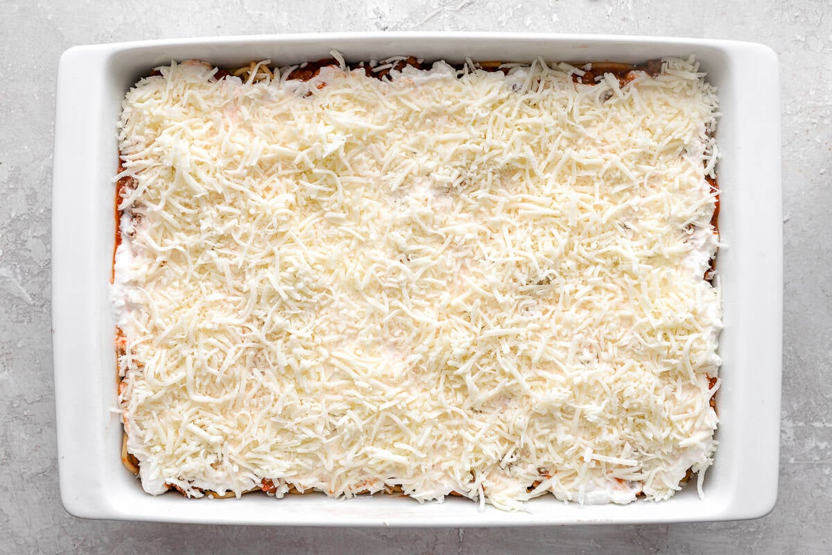A white casserole dish filled with unbaked million dollar spaghetti, topped with a thick layer of shredded mozzarella cheese over a creamy cheese mixture.