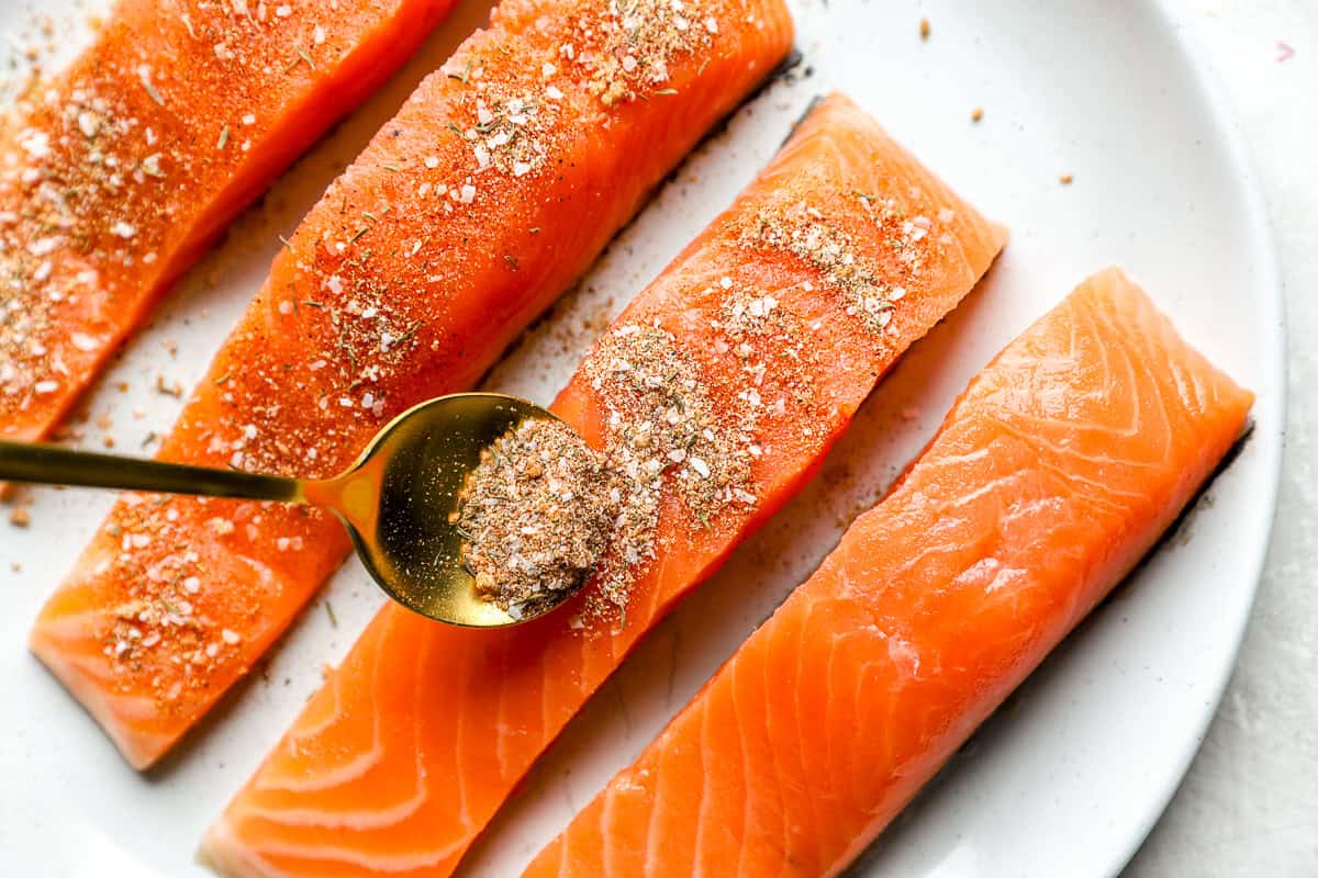 Raw salmon fillets on a plate being sprinkled with homemade seasoning using a gold spoon.