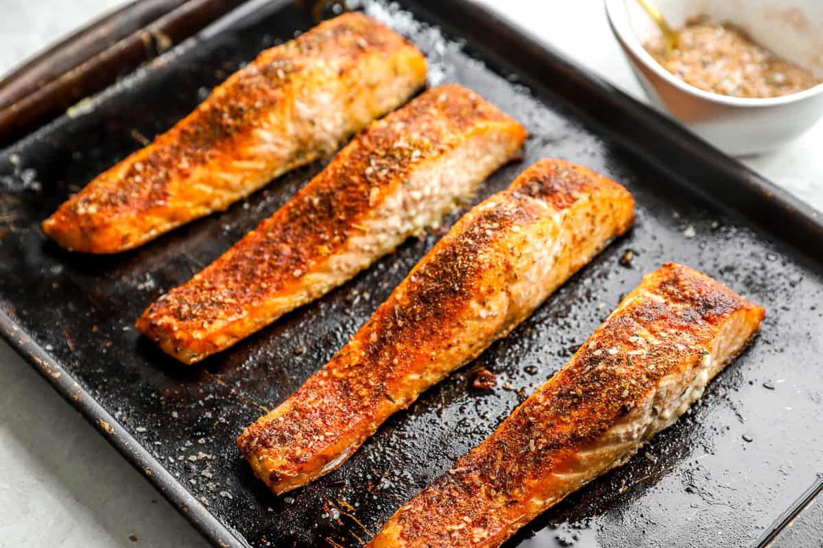 Four seasoned salmon fillets freshly baked on a dark sheet pan, golden and crisp on the edges.